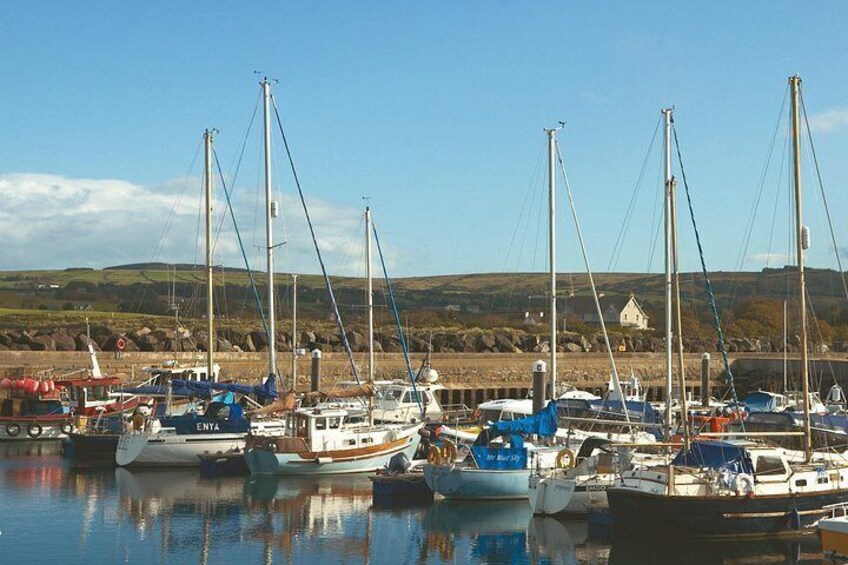 A marina with several sailboats docked, a small house in the background, and a hilly landscape. 2