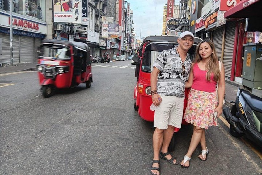 A street with tuk-tuks, a red auto-rickshaw, and a man and woman standing in front of a shop. 1