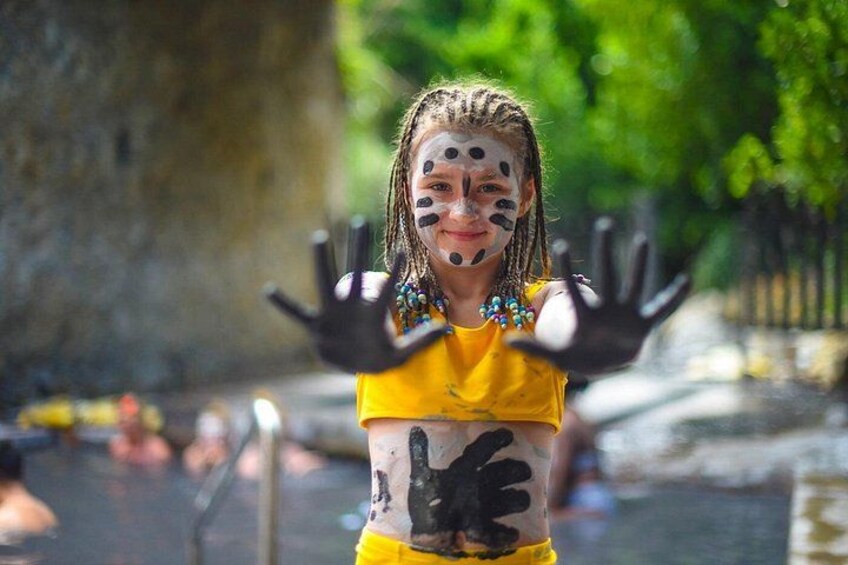 A person in a yellow top with black paint on their hands and face, standing by a body of water. 1