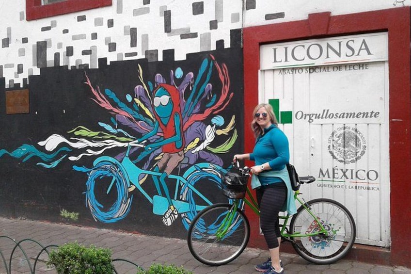 Een muurschildering van een kleurrijke fiets, een vrouw die naast een groene fiets staat, en een gebouw met een bord waarop 'LICONSA' staat. 1