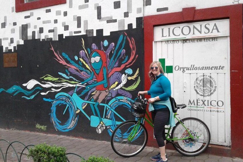 Un mural con una bicicleta de colores en una pared, una mujer de pie junto a una bicicleta verde, y un edificio con un letrero que dice 'LICONSA'. 1