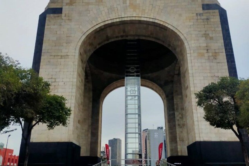Une grande arche en pierre, avec une structure métallique au centre, entourée d’arbres et d’immeubles urbains. 2