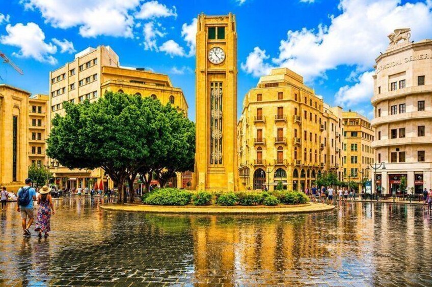 A historic square with a prominent clock tower, surrounded by classical architecture and a central fountain reflecting the sky. 1