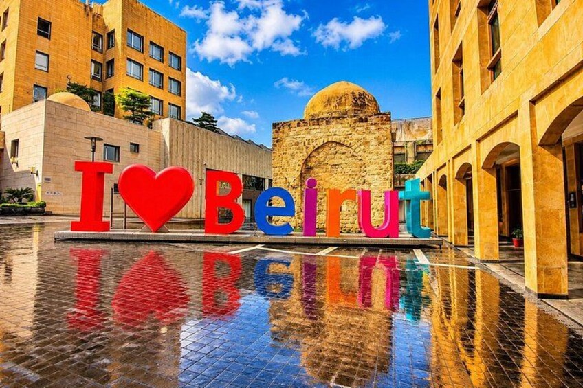 A reflective water surface with the words 'I ♥ Beirut' in colorful letters, a stone archway, and modern buildings in the background. 2