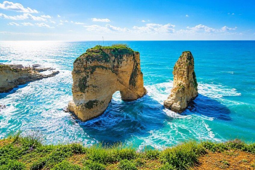 A coastal landscape with a prominent rock formation arching over the sea, surrounded by clear blue waters and a clear sky. 4