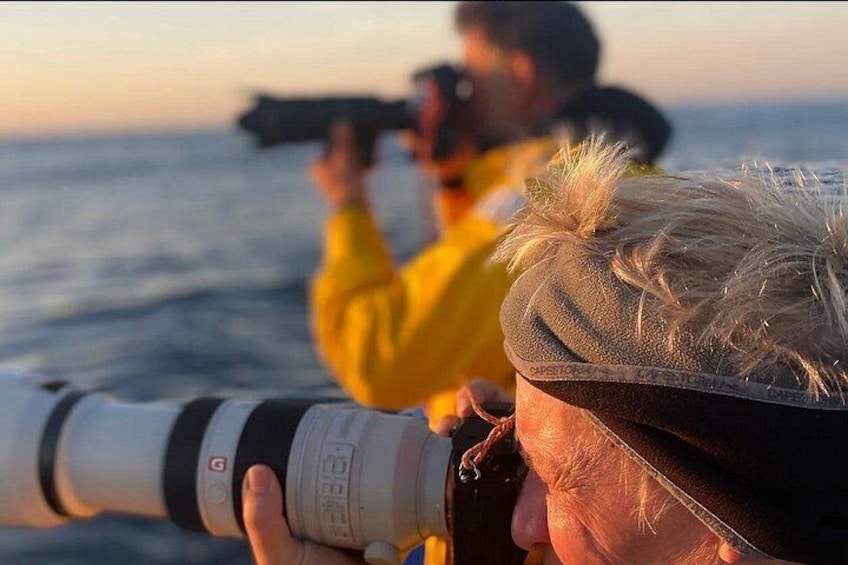 A person holding a camera with a strap, another person in the background with a camera, and a body of water. 3