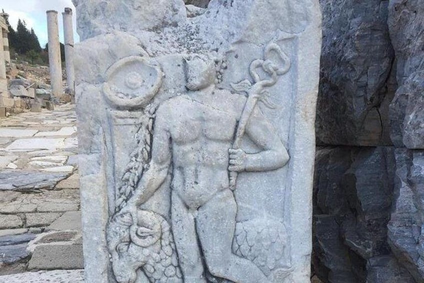 En stenhugget figur med et trident og et skjold, med en slange snoet rundt om armen, foran en stenet baggrund. 5