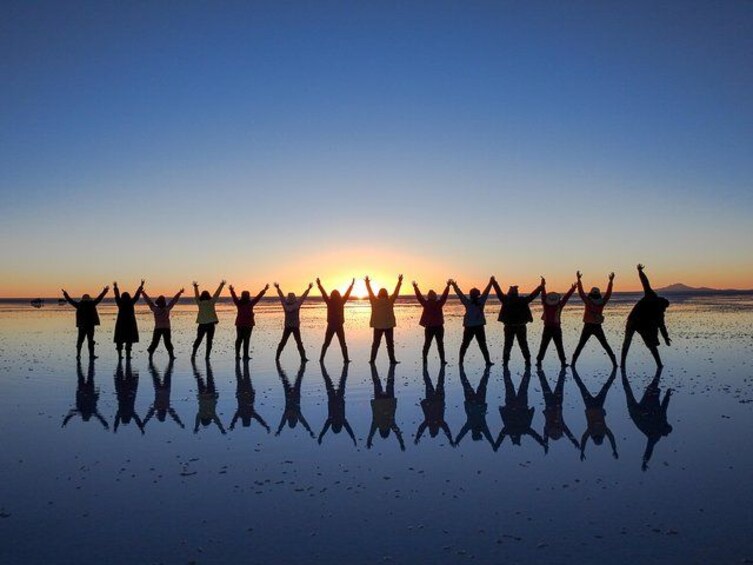 A group of people silhouetted against a sunset on a reflective surface, likely a beach. 1