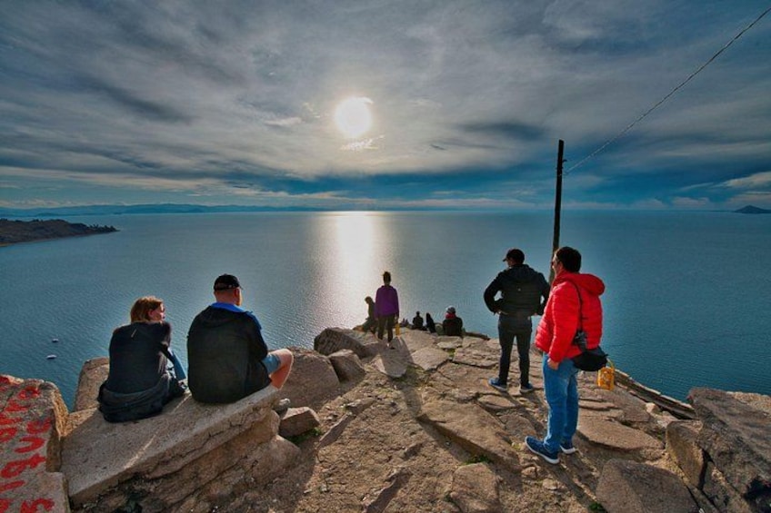 Eine Gruppe von Menschen auf einer felsigen Aussichtsplattform an einem ruhigen Gewässer, mit untergehender Sonne im Hintergrund. 4