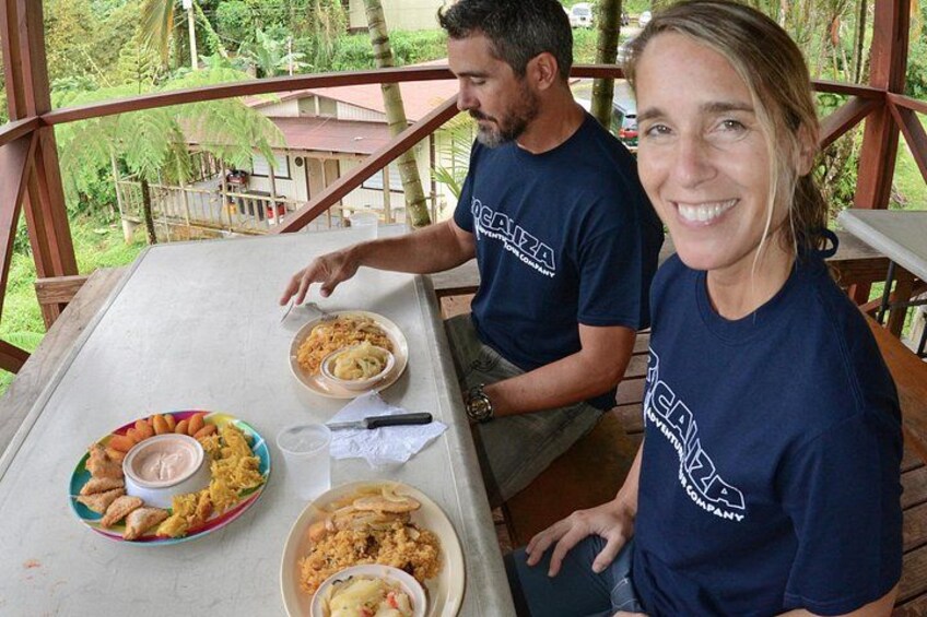 Two people sitting at a table with food, outdoors with a wooden deck and greenery in the background. 2