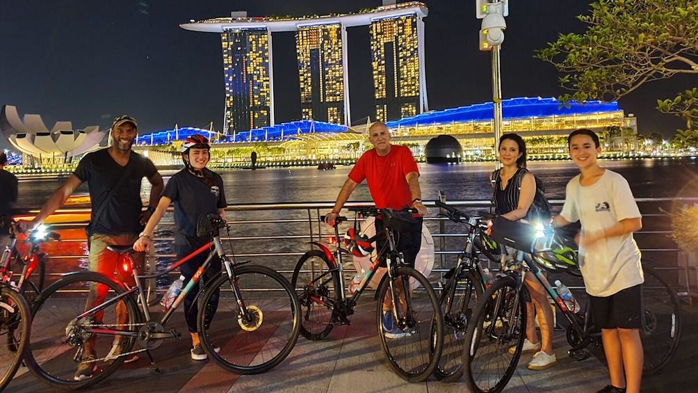 A group of people standing with bicycles in front of a brightly lit waterfront with a distinctive building in the background. 5