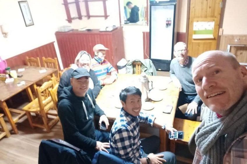 A group of people seated around a wooden dining table in a room with wooden walls and a refrigerator in the background. 5
