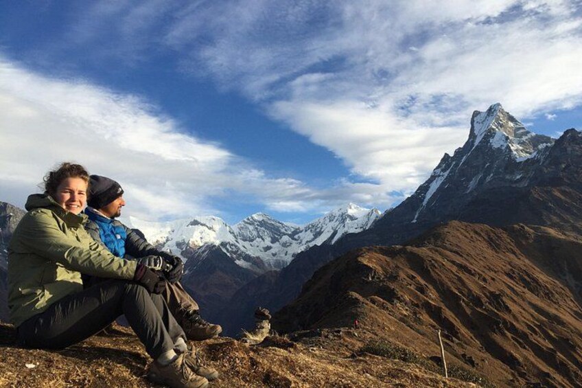 Two people sitting on a mountain ridge with snow-capped peaks in the background. 1