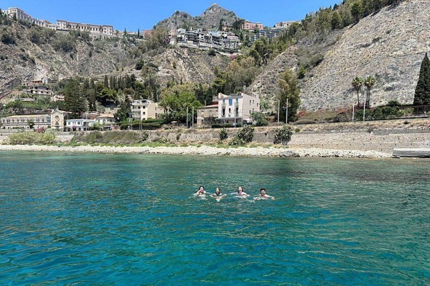Un groupe de personnes nage dans une mer d’un bleu limpide, avec en arrière-plan des montagnes et des bâtiments. 3