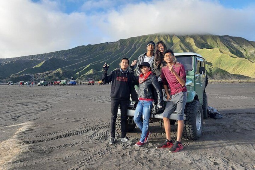 A group of people posing in front of a green vehicle on a sandy beach with mountains in the background. 4