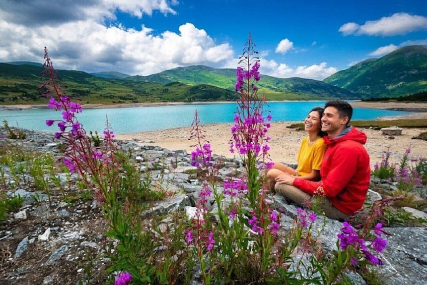 A couple sitting on a rocky shore with purple flowers and a lake in the background. 1