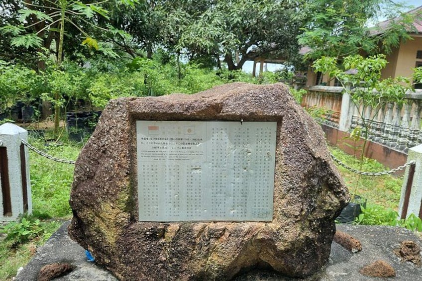 A large stone with a plaque containing text, surrounded by greenery and a fence. 3