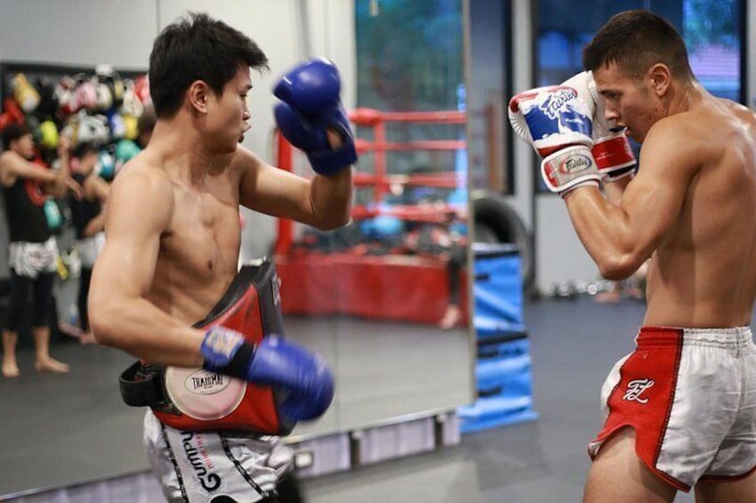 Two individuals engaged in a boxing match in a gym with boxing equipment and other people in the background. 4