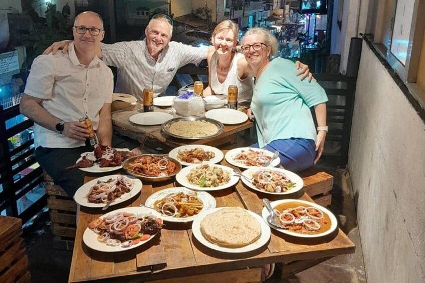 A group of people around a wooden table with various dishes, including pasta and bread, in an outdoor setting. 3