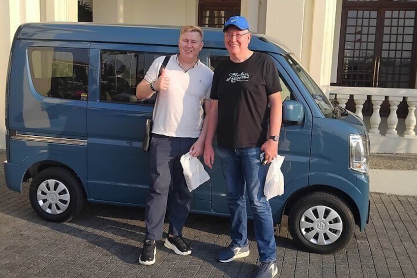 Zwei Personen stehen neben einem blauen Lieferwagen, im Hintergrund ein Gebäude. 1