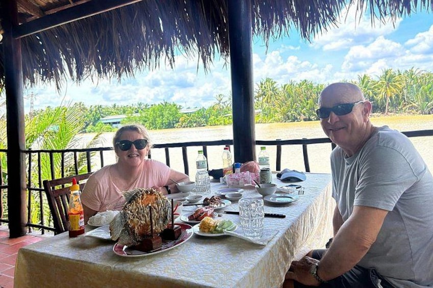 Twee personen dineren aan een buitentafel met uitzicht op een rivier en palmbomen. 3