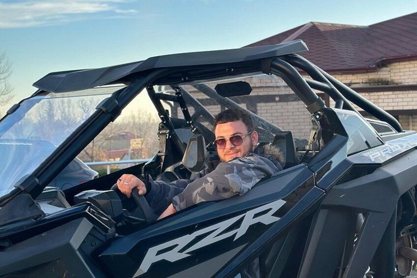 A person in a black Polaris RZR vehicle with a roof, parked outdoors. 3