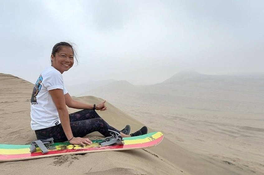 A person sitting on a sand dune with a colorful board, surrounded by a vast sandy landscape and distant mountains. 2