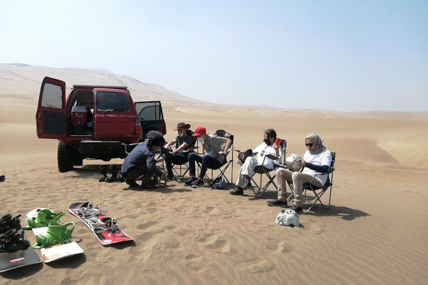 A group of people sitting on chairs in a desert with a red vehicle in the background. 4