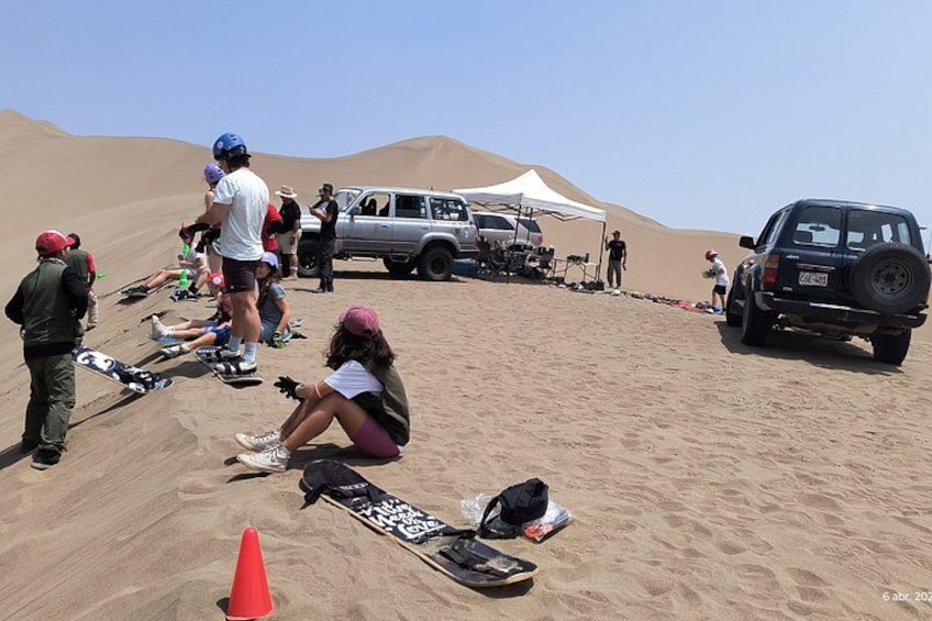 A group of people sitting on a sandy hill with a tent and vehicles in the background. 3