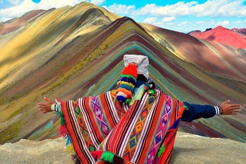 A person in colorful traditional attire standing on a mountain ridge with arms outstretched. 3