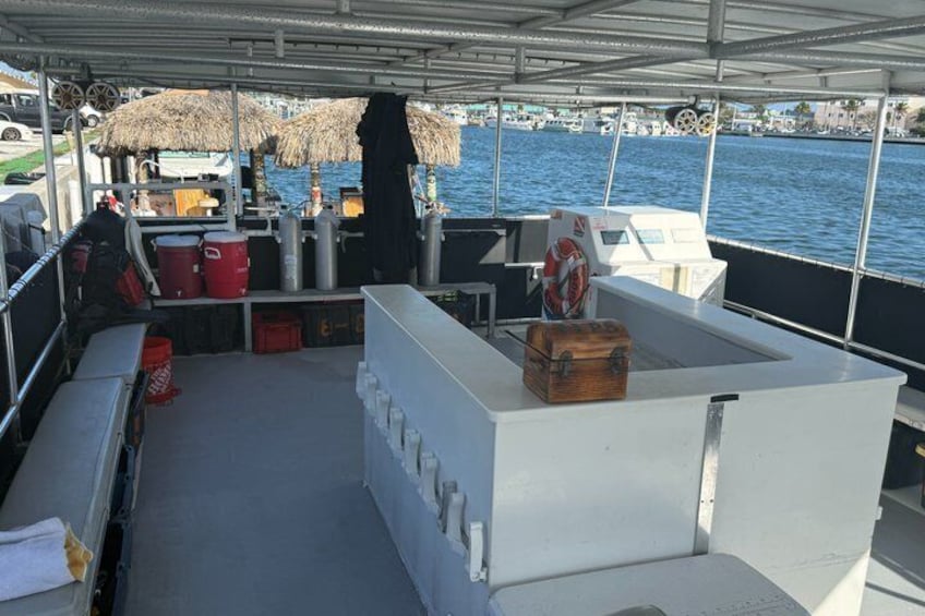 Cubierta de barco con zona de bar blanca, barriles rojos, un flotador salvavidas y una sombrilla de paja. 3