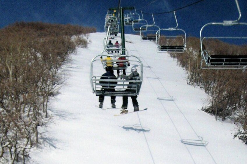 A ski lift chair ascending a snowy slope with bare trees on either side. 1