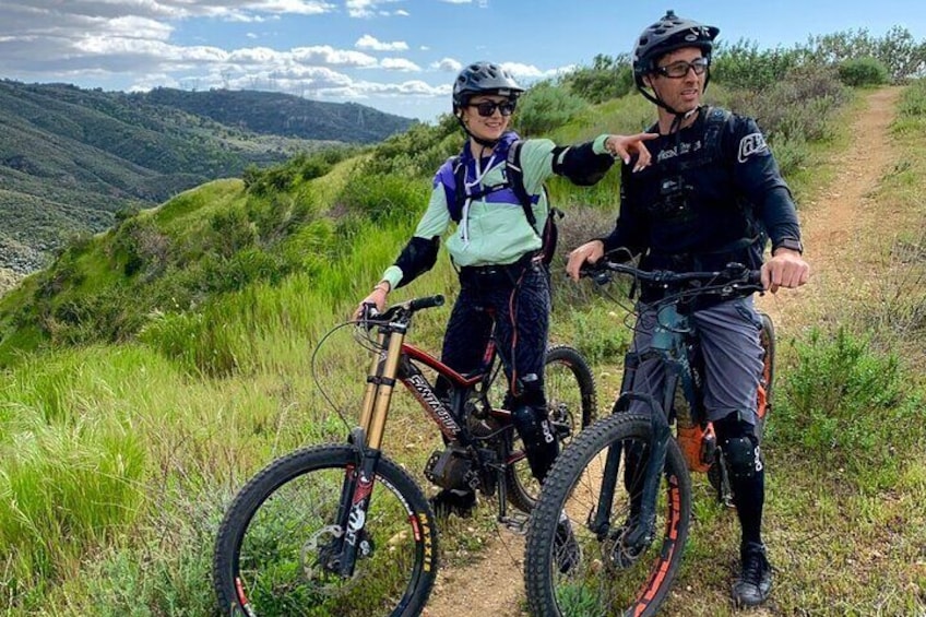 Two people on a mountain biking trail with a scenic view of hills and a clear sky. 1