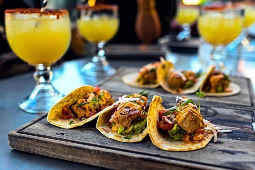 Tacos on a wooden board with drinks in the background. 1