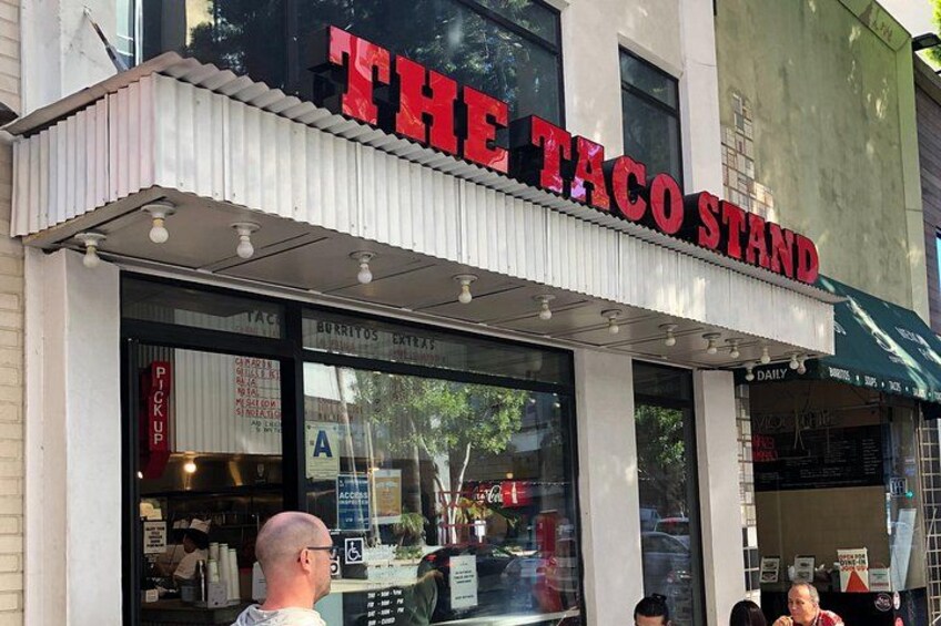 A taco stand with a red sign, large windows, and a person walking by. 2