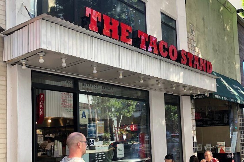A taco stand with a red sign, a window displaying menu items, and a person standing outside. 2