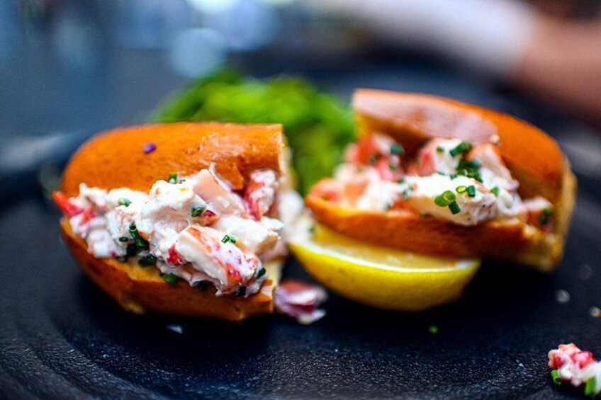 A lobster roll with a lemon wedge and green garnish on a dark plate. 4