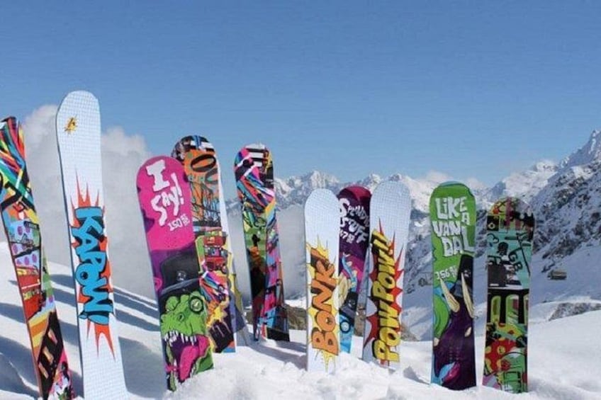 Eine Reihe bunter Snowboards mit verschiedenen Designs und Schriftzügen vor einer verschneiten Bergkulisse. 1