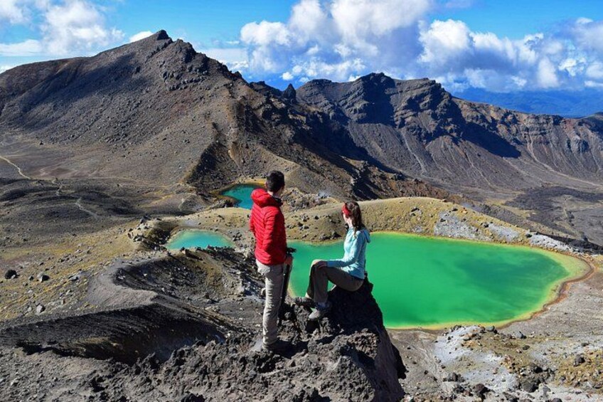 Two people standing on a rocky outcrop overlooking green lakes and mountains. 1