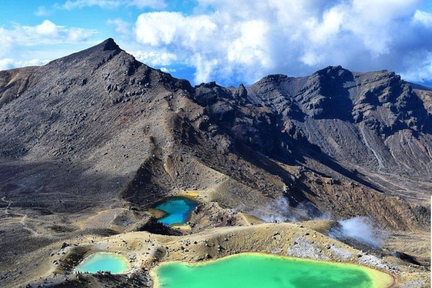 A mountainous landscape with a prominent peak, several small lakes with varying colors, and steam rising from the ground. 2