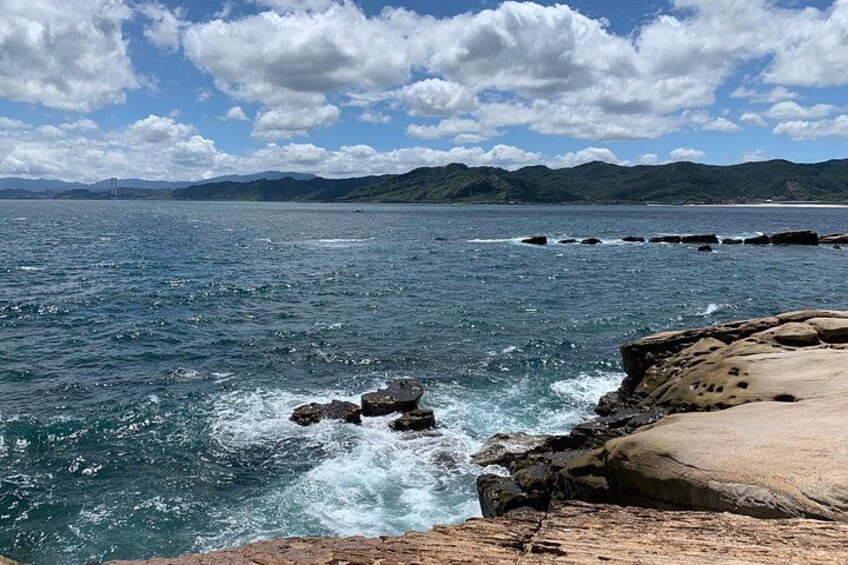 A coastal landscape with rocky shores, waves crashing against the rocks, and mountains in the distance under a partly cloudy sky. 2