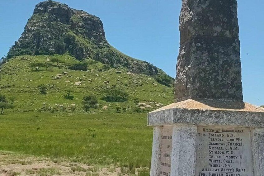 Et monument med en liste over navne, foran en græsklædt bakke og et klippefremspring. 2