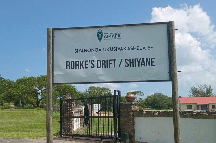 Et skilt med teksten 'SIYABONGA UKUSIVAK ASHELA E- RORKE'S DRIFT / SHIYANE' og en låge. 5
