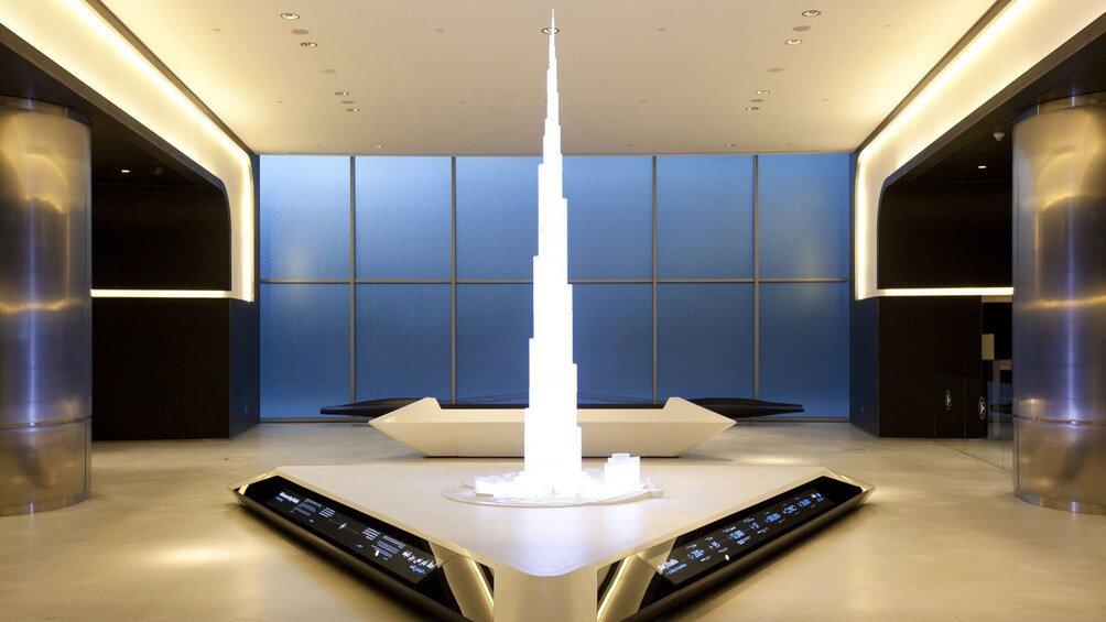 Een modern interieur met een centrale witte constructie die op een torenspits lijkt, omringd door slanke, metalen kolommen en een blauwachtige muur. 3