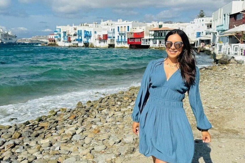 A person standing on a rocky beach with white buildings and a red boat in the background. 5