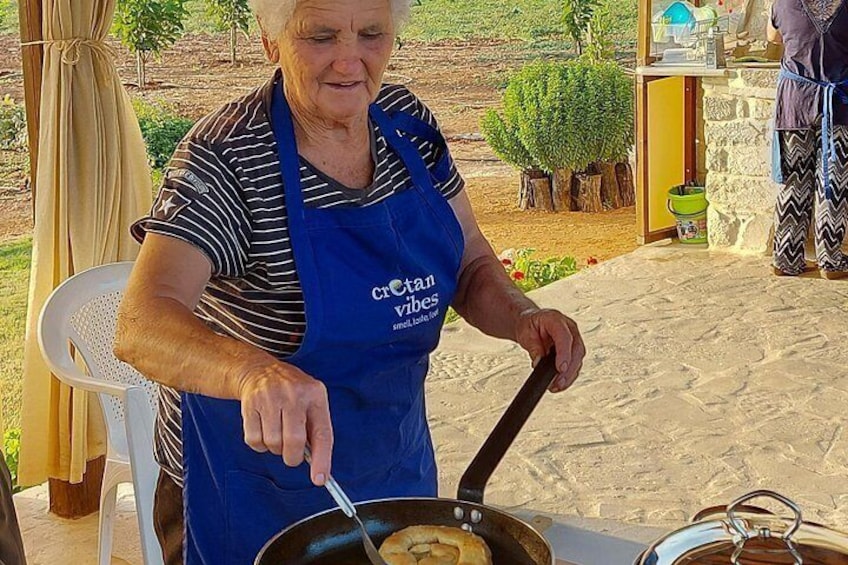 An elderly woman cooking outdoors, wearing a blue apron, with a pot on a stove and a chair nearby. 1
