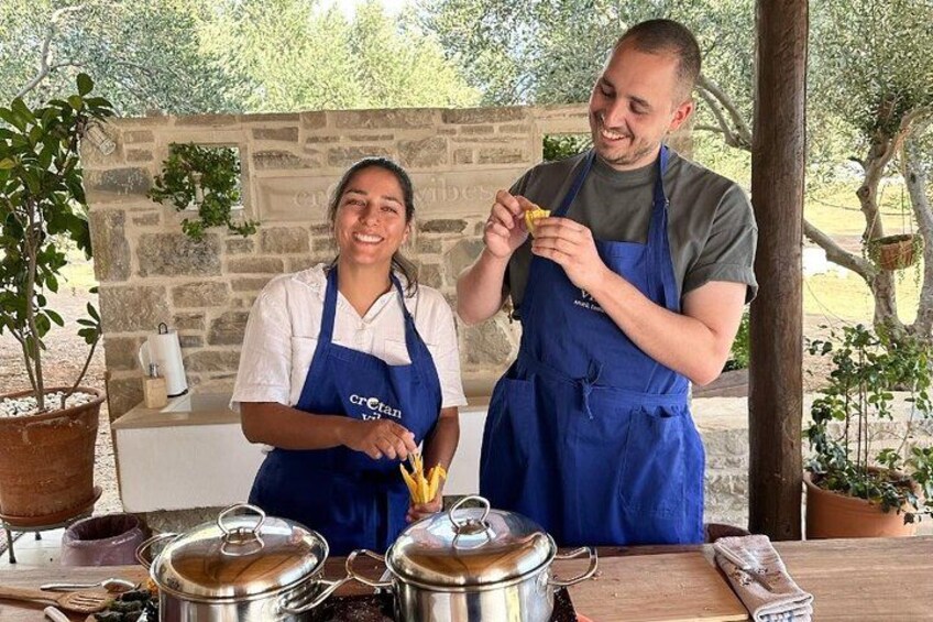 Two people in aprons preparing food outdoors with cooking equipment and plants in the background. 4