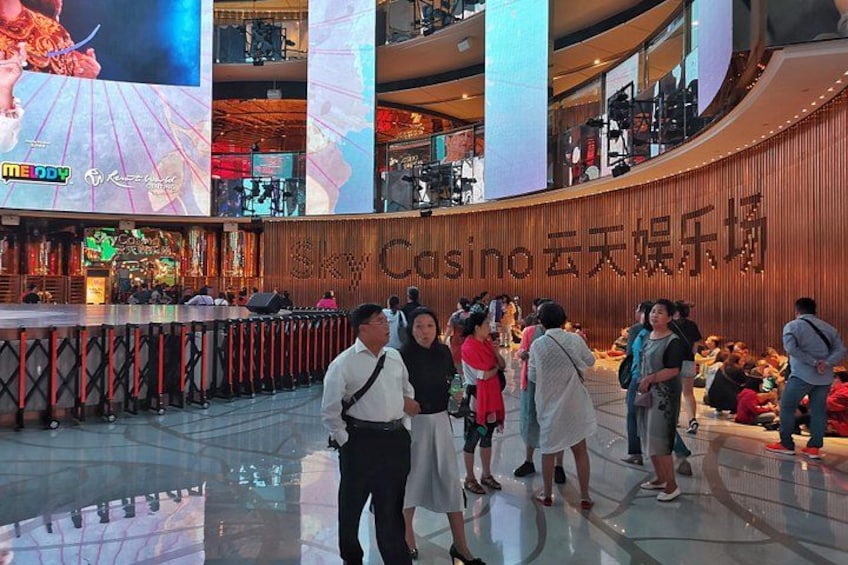 Interno di un casinò con un grande schermo digitale che mostra un gioco, una parete in legno con il nome del casinò e persone che passeggiano. 4