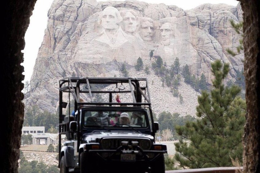 A vehicle with passengers, a large rock formation with carved faces, and a tree in the foreground. 2