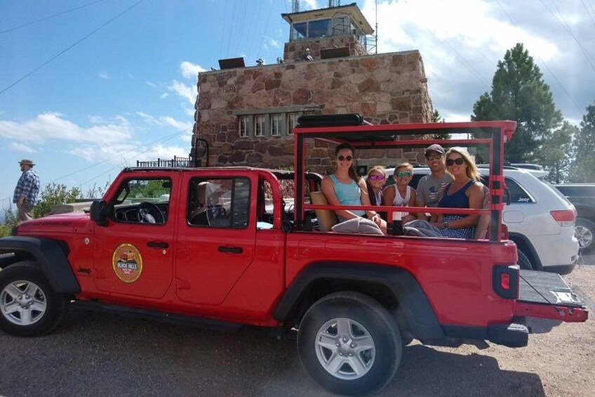 A red Jeep with passengers, parked in front of a stone building. 1